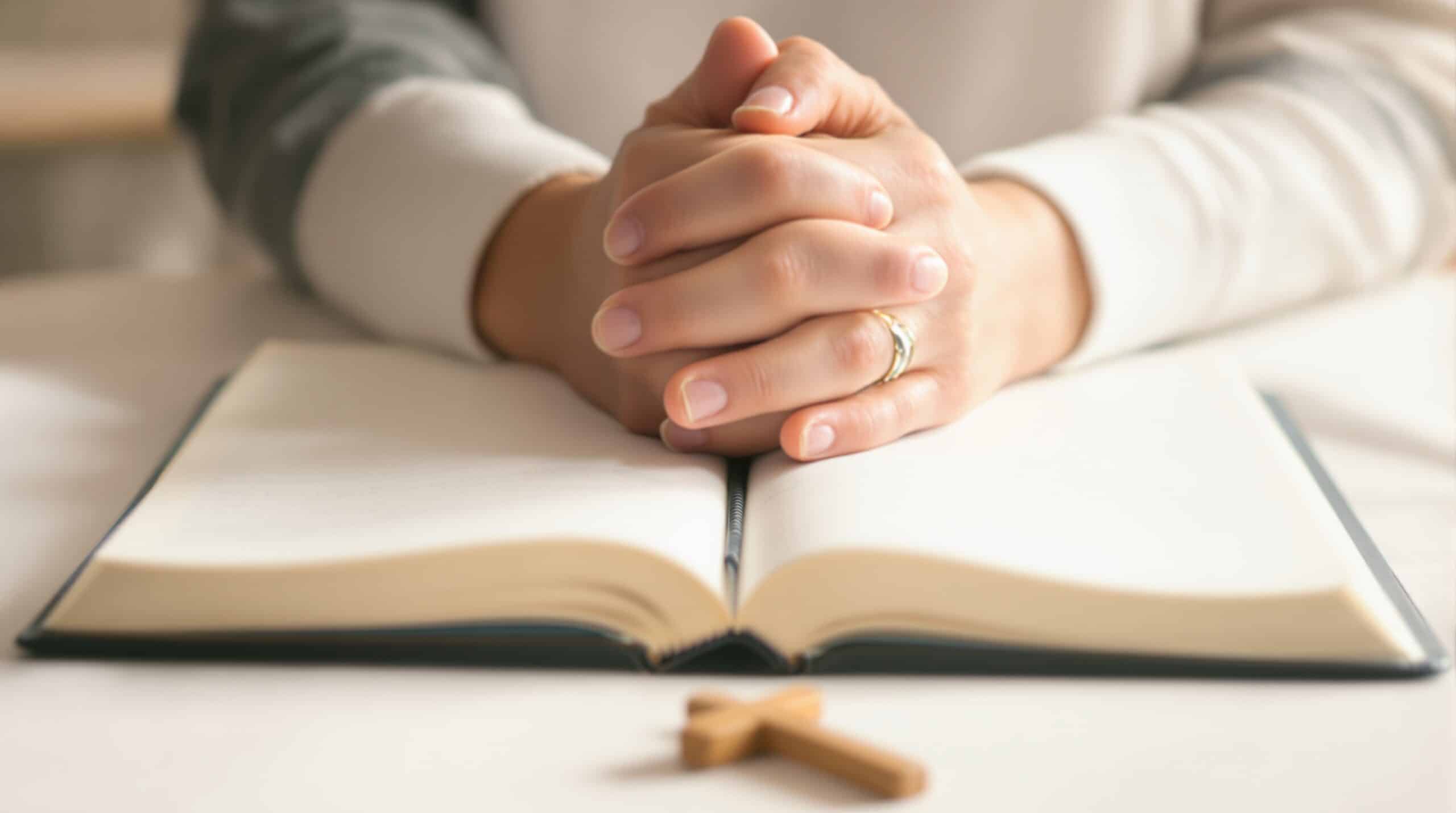 Hands clasped in prayer above open journal with wooden cross nearby, soft natural lighting creating peaceful atmosphere