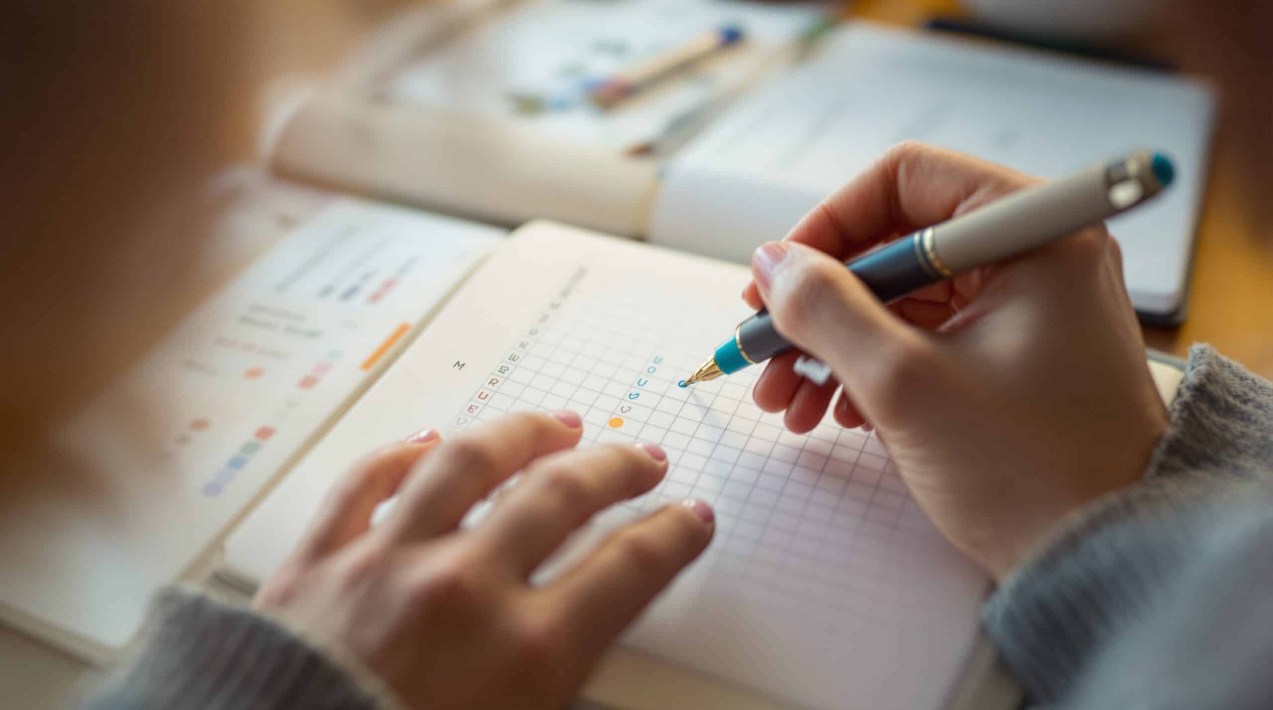 Close-up of hands using colored pen to mark completed habit in bullet journal grid with checkmarks and tracking dots
