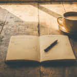 An open journal with writing prompts on a sunlit desk with a pen and coffee cup, creating a peaceful morning journaling scene.