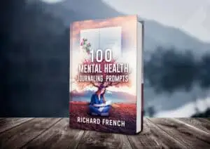 Alt text: "Book cover mockup of '100 Mental Health Journaling Prompts' by Richard French, featuring a woman sitting cross-legged while writing in a journal with artistic tree branches extending from her head, set against a desert landscape with floating journal pages, displayed on wooden planks with a blurred mountain background.